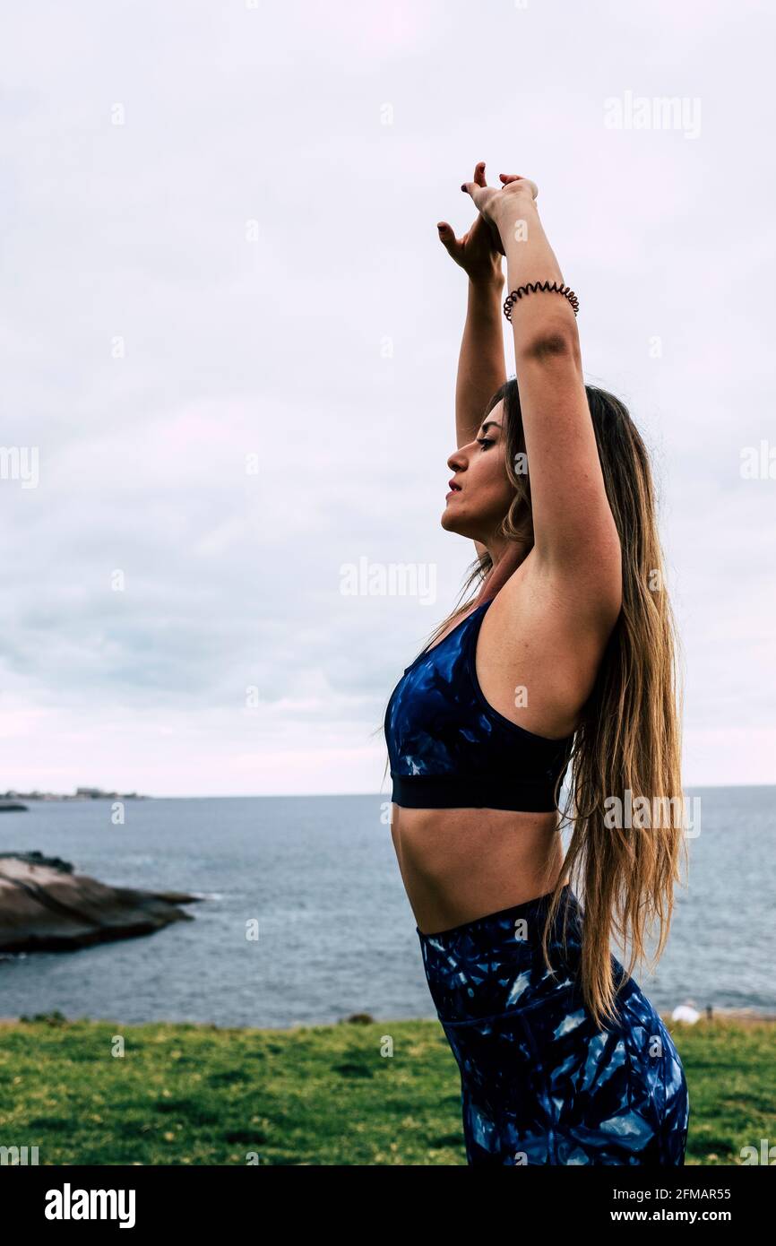 Giovane donna adulta in piedi fare stretching pilates esercizi di yoga equilibrati per la cura del corpo sport tempo libero attivo - persone sane in allenamento giorno di svago con oceano in background Foto Stock