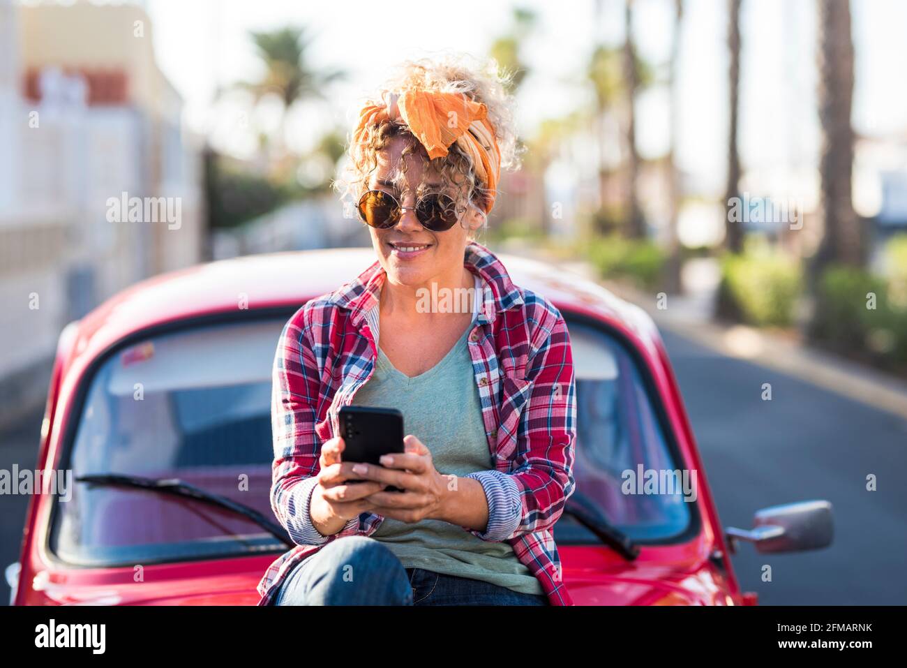Donna caucasica giovane adulta alla moda usa una moderna connessione telefonica esterno vicino alla sua bella auto rossa in viaggio e indipendente stile di vita - concetto di messaggistica app e chat di testo Foto Stock