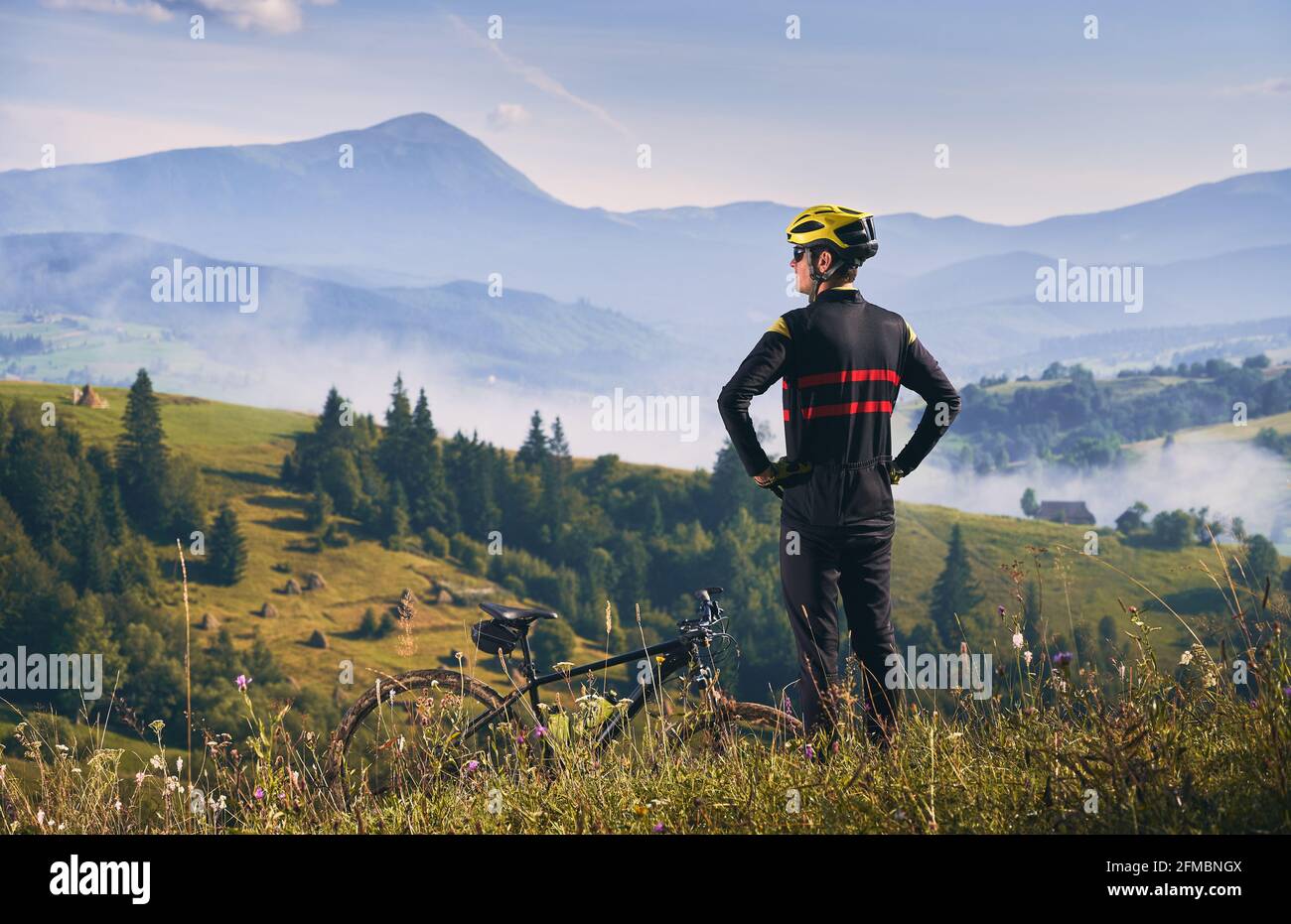 Vista posteriore su un ciclista che indossa uniforme e casco, in piedi su una collina che guarda il bellissimo paesaggio delle montagne. La mattina presto nelle montagne soleggiate e nebbose. Concetto di unità con la natura Foto Stock