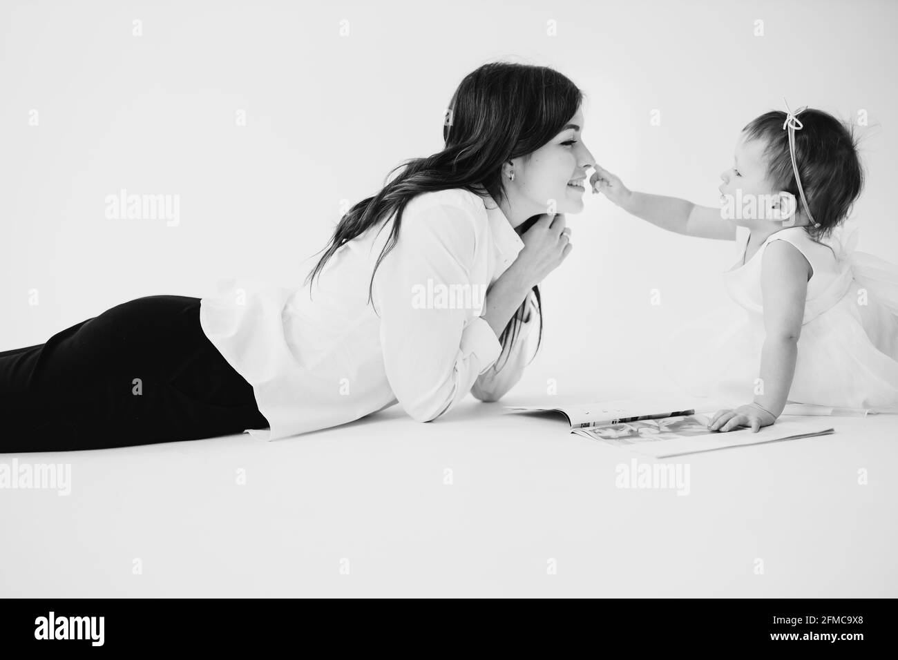 Foto in bianco e nero di mamma felice che si posa sul pavimento con la figlia del bambino e che legge la rivista, bambina che tocca delicatamente il naso della madre Foto Stock