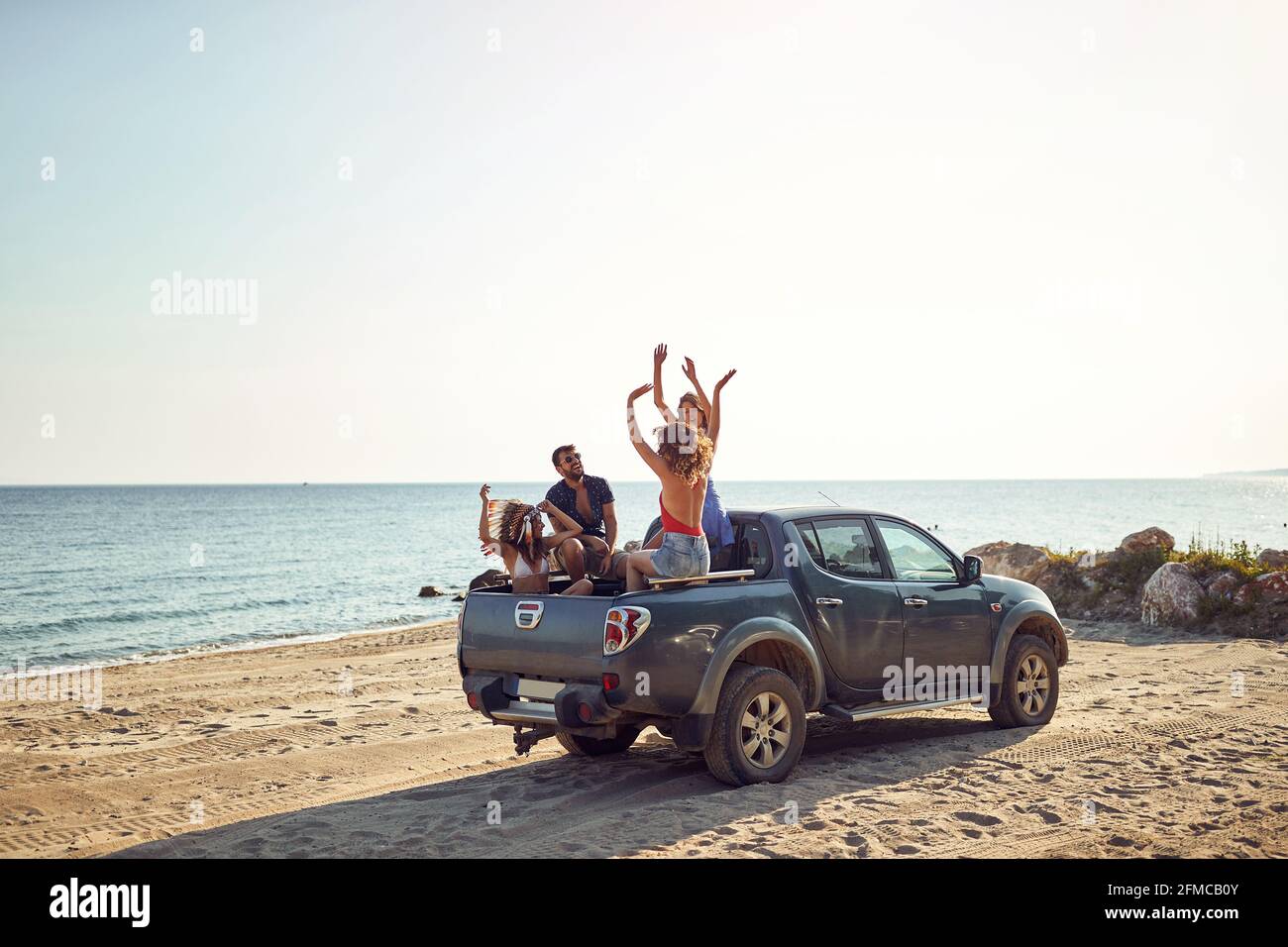 Un gruppo di bei modelli sta ballando e gustando la musica sul retro di un'auto in riva al mare in una bella giornata di sole. Estate, mare, vacanza, amicizia Foto Stock
