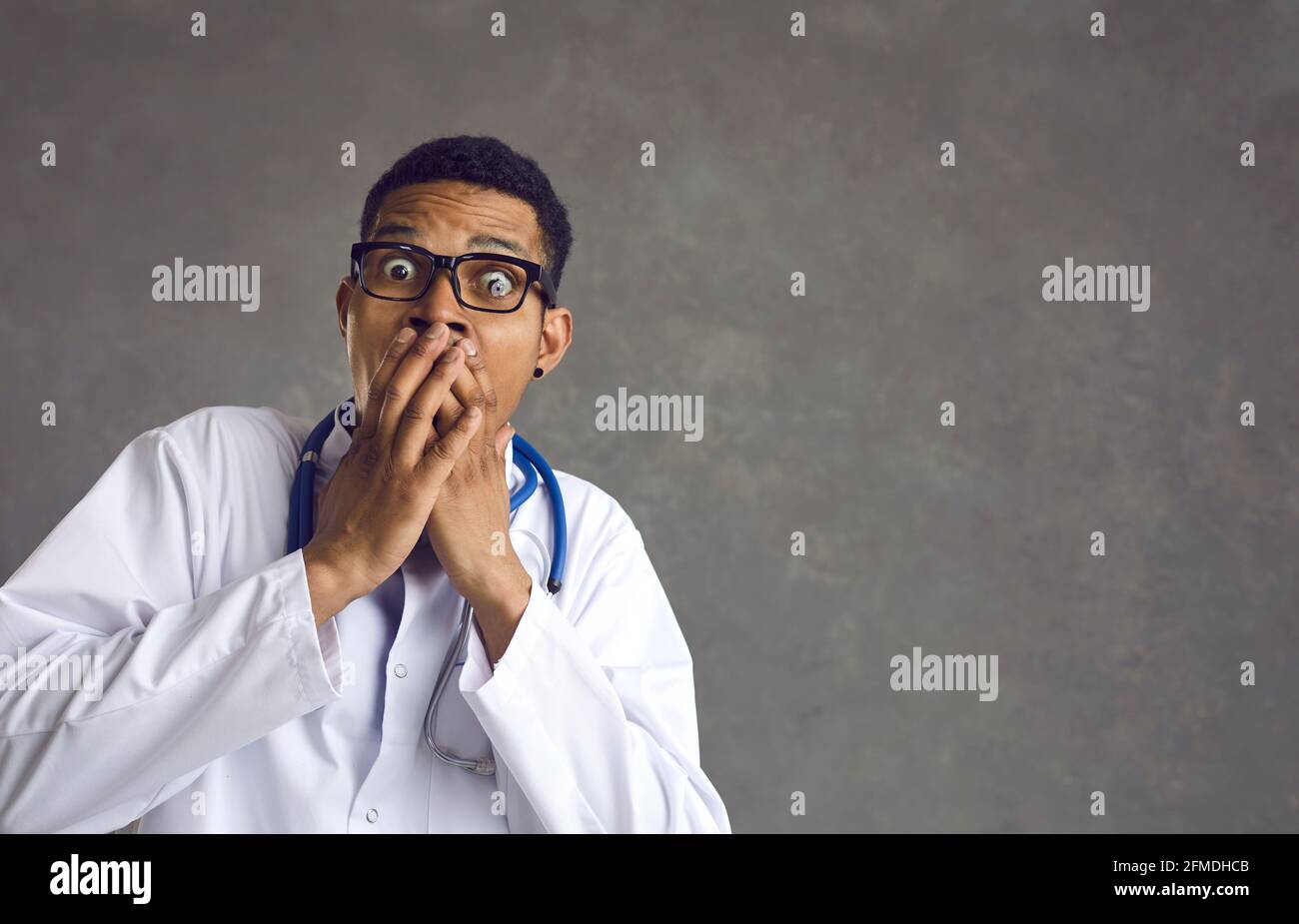 Il medico africano americano maschio con un'espressione spaventata copre la sua bocca su uno sfondo grigio. Foto Stock