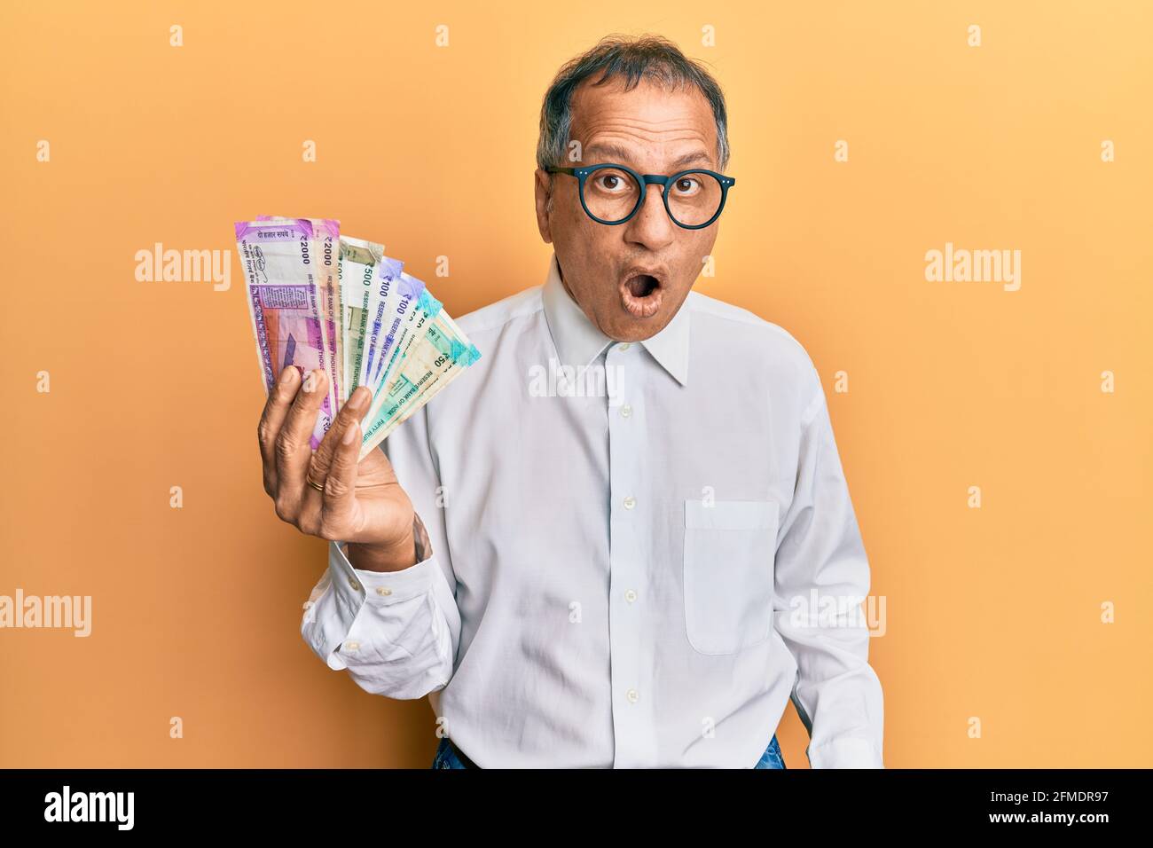 Uomo indiano di mezza età che tiene le banconote di rupia indiane spaventato e stupito con la bocca aperta per sorpresa, faccia di incredulità Foto Stock