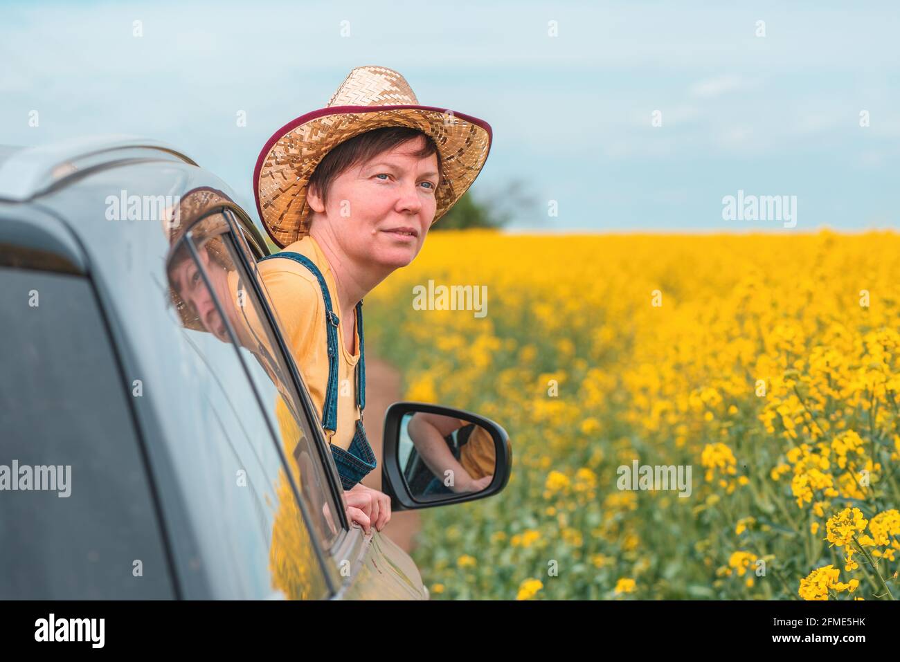 Donna che ama viaggiare in auto in un paesaggio fiorente della campagna estiva, testa con cappello di paglia che raggiunge il finestrino, fuoco selettivo Foto Stock