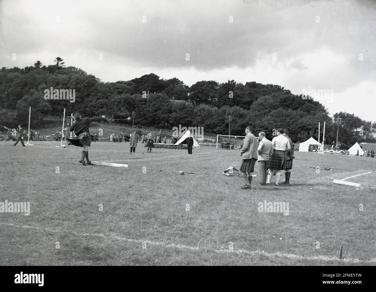 1956, storico, fuori in un campo d'erba a un highland giochi, un maschio concorrente in un kilt, che prende parte alla gara di lancio del martello, guardato da funzionari, Scozia, Regno Unito. Il martello è fatto da una sfera di metallo sferica fissata ad un manico, che è usato per girare il martello intorno alla loro testa e poi per gettarlo il più possibile, la distanza più grande essendo il vincitore. Nei giochi Highland non ci sono margini al contrario del lancio del martello nei Giochi Olimpici. Foto Stock