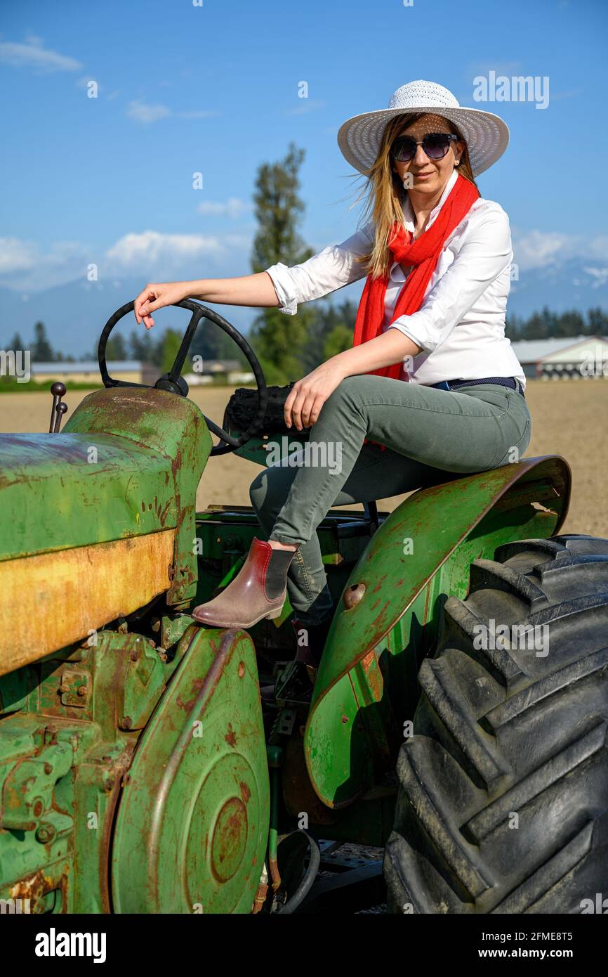 Donna urbana in abbigliamento casual seduta su un vecchio trattore contro il cielo blu nel terreno agricolo Foto Stock