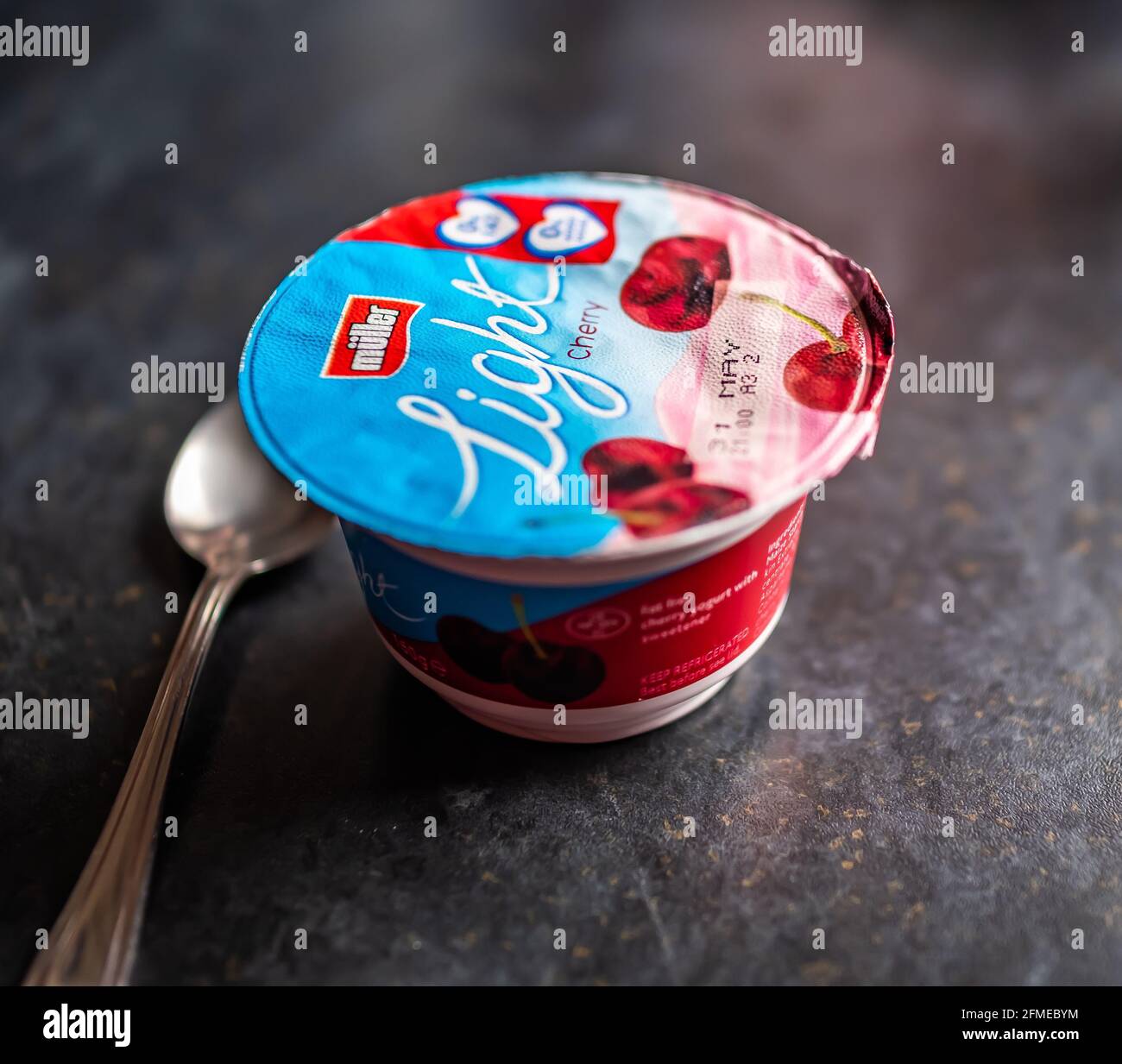 Primo piano e fuoco selettivo su una vasca di Muller Yogurt alla amarena senza grassi e un cucchiaio d'argento su un piano di lavoro nero Foto Stock