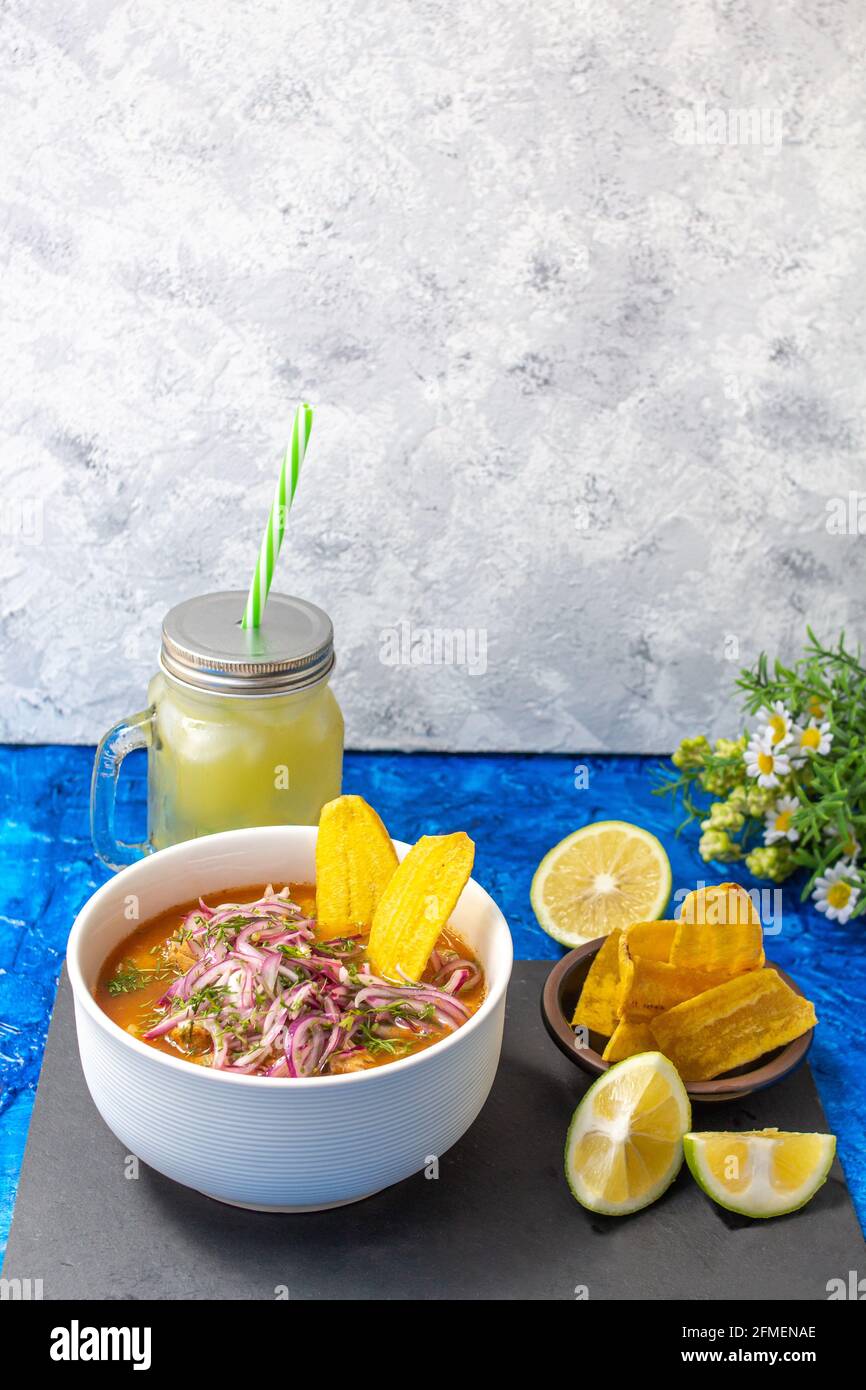 Piatto di encobollado, un alimento tipico dell'Ecuador preparato con pesce, cipolla e yucca con una caraffa di limonata e scaglie di banana su un blu e una ruggine Foto Stock