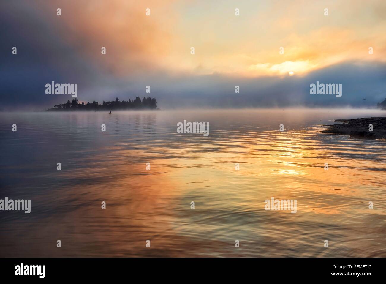 Il sole sorge sull'orizzonte dell'acqua sul lago Jindabyne nelle Montagne Snowy australiane - luce solare panoramica in condizioni di nebbia nebbiosa. Foto Stock