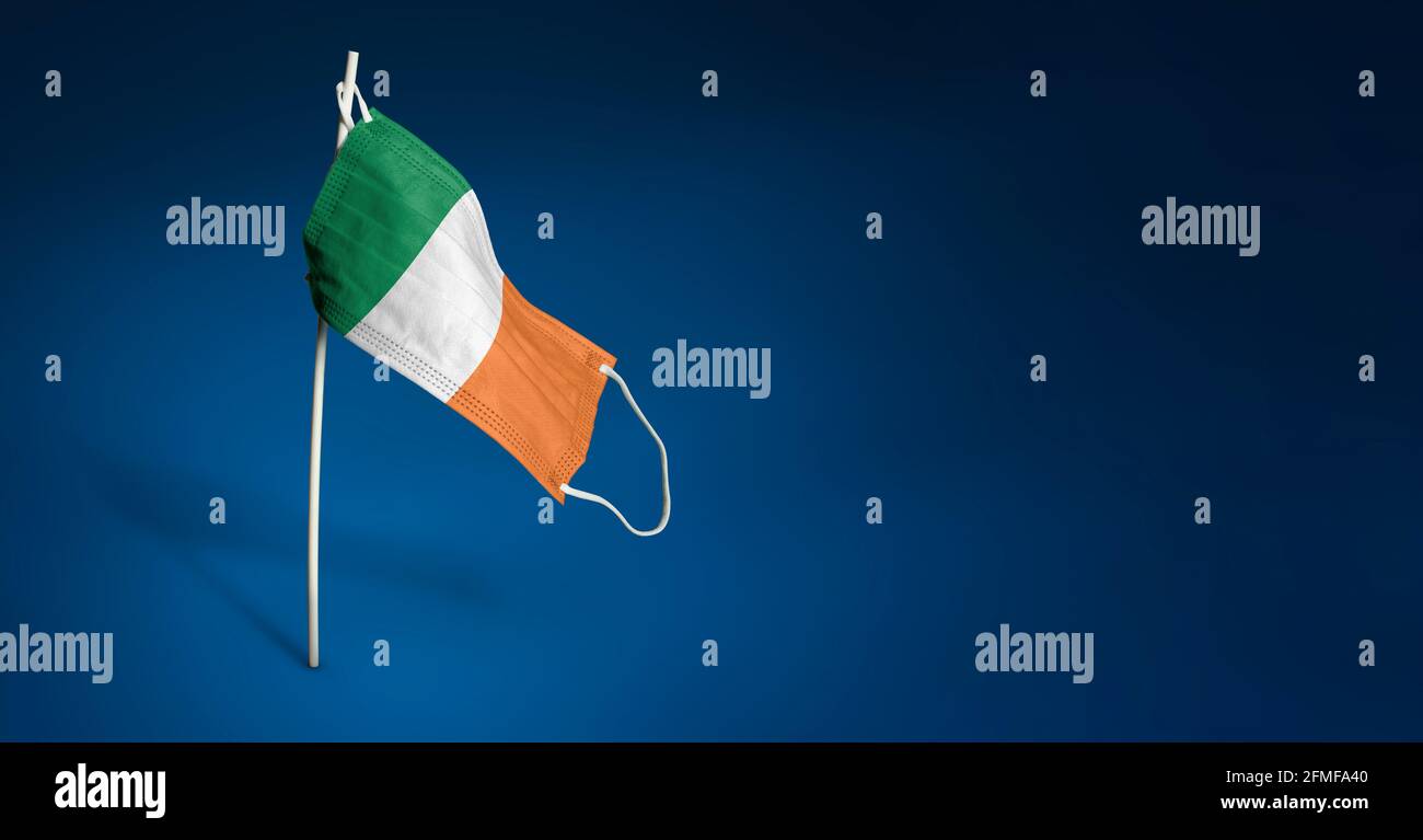 Maschera irlandese su sfondo blu scuro. Bandiera ondulata dell'Irlanda dipinta su maschera medica sul palo. Flag di attacco del virus. Concetto del banner della Fight ag Foto Stock