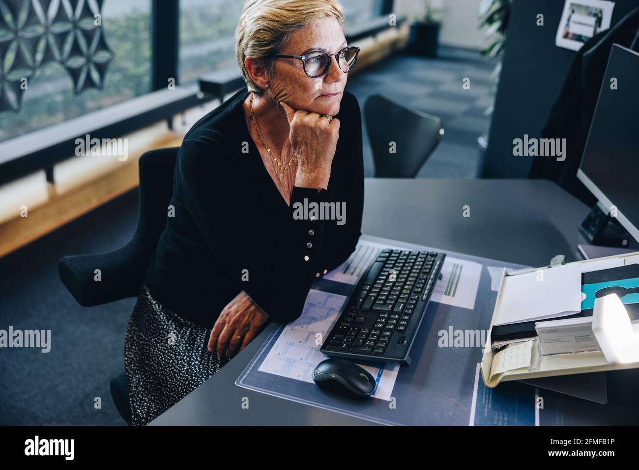 Donna d'affari anziana seduto alla sua scrivania guardando il computer. Donna con la mano sul mento al suo posto di lavoro. Foto Stock