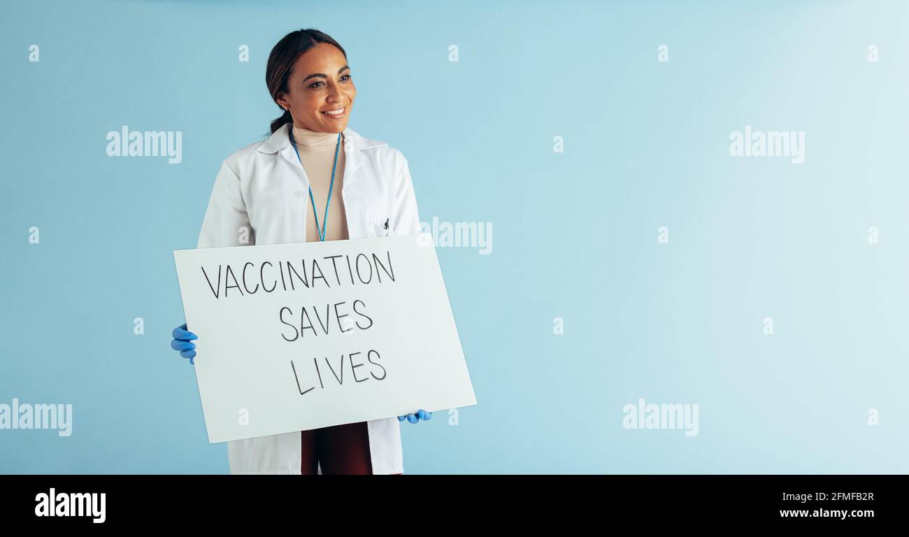 Medico che guarda via e sorridendo con il banner 'vaccinazioni salva vitea' su sfondo blu. Operatore sanitario che mostra un segno di consapevolezza della vaccinazione Foto Stock
