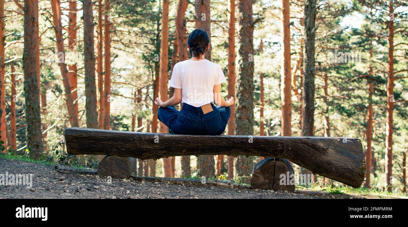 Una giovane donna seduta sulla schiena su una panchina mentre fa yoga nella foresta. Orizzontale. Foto Stock
