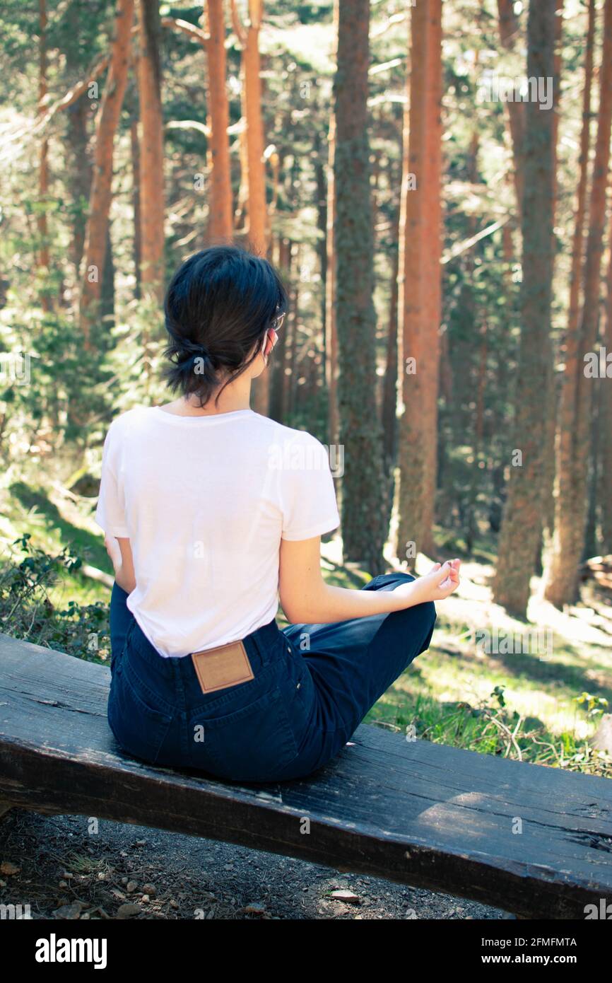 Giovane donna seduta su una panchina mentre medita in una foresta di alberi alti. Verticale. Foto Stock