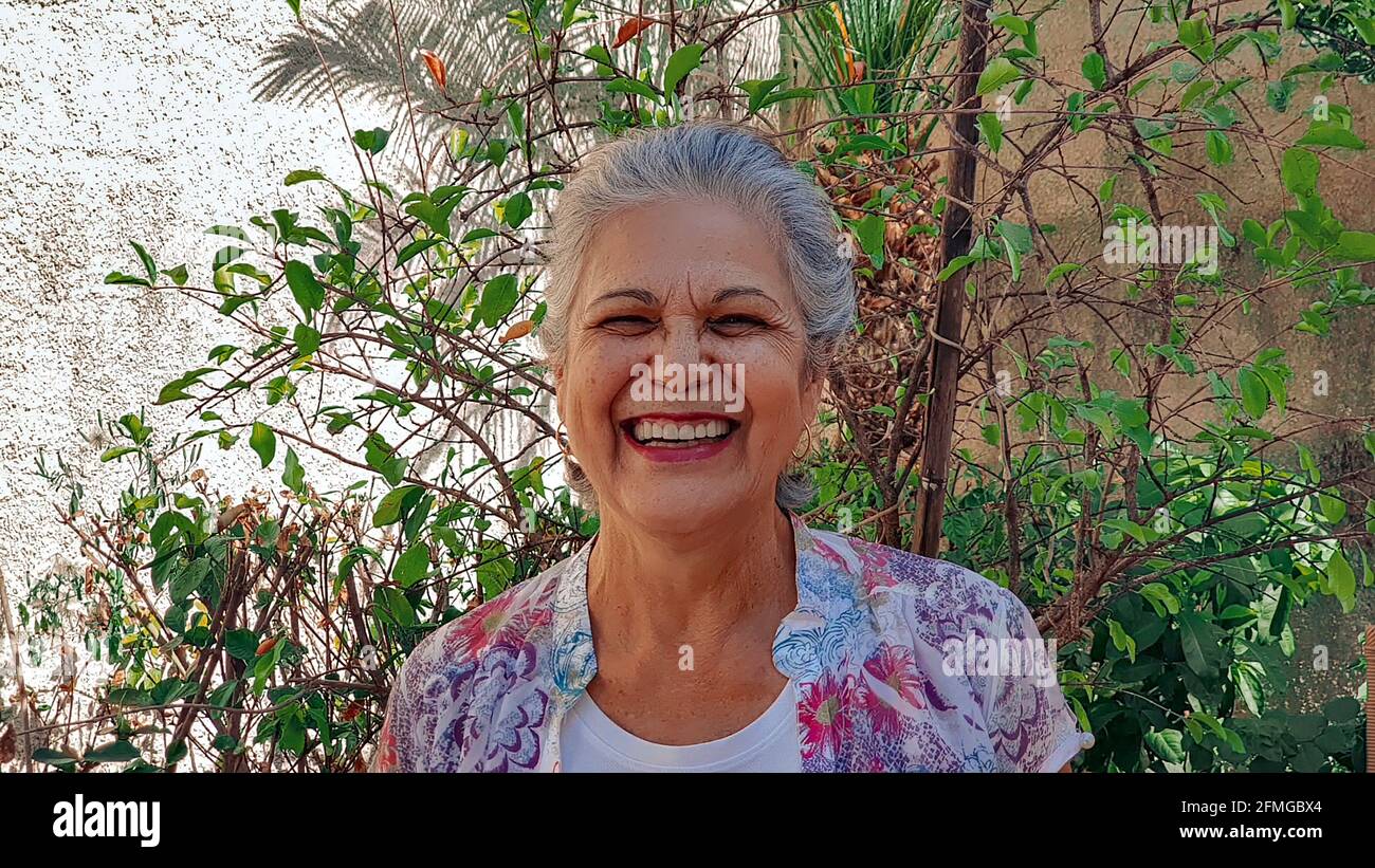 Donna matura che sorride nella casa del cortile con sole e piante. Donna bianca con capelli grigi a casa. Foto Stock