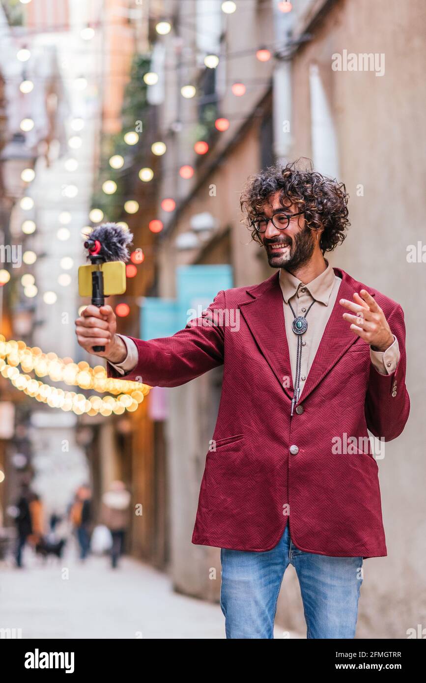 Attraente uomo caucasico che si registra nelle strade di Barcellona. Sta tenendo un gimbal con un telefono cellulare giallo e un microfono. Sta vestendo casual con jeans blu e giacca rossa. Foto Stock