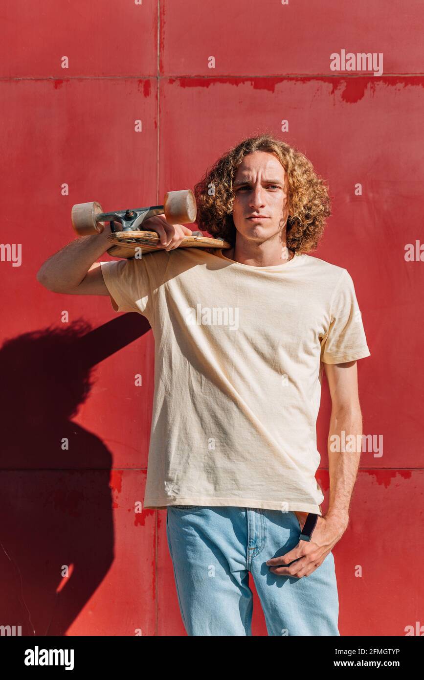 ritratto verticale di un giovane che porta uno skateboard su questa spalla. Ha lunghi capelli ricci rossi e abiti casual. Sfondo rosso con ombre profonde Foto Stock