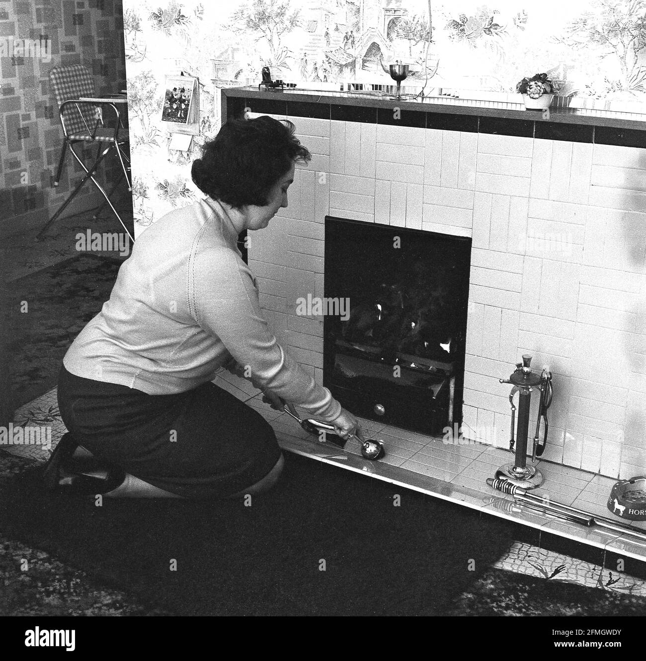 anni '60, storico, all'interno di un soggiorno, una signora che starnutisce a un fuoco di carbone, la pulizia delle piastrelle di fronte al fuoco. Foto Stock