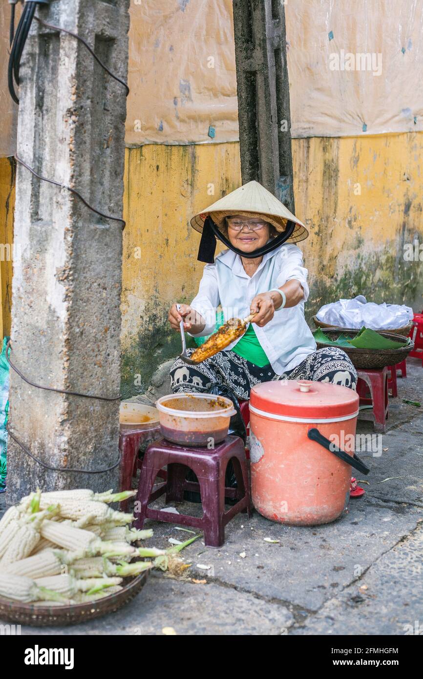 Anziana venditrice di strada vietnamita che vende spuntini dal Pavement Cafe, Hoi An, Vietnam Foto Stock