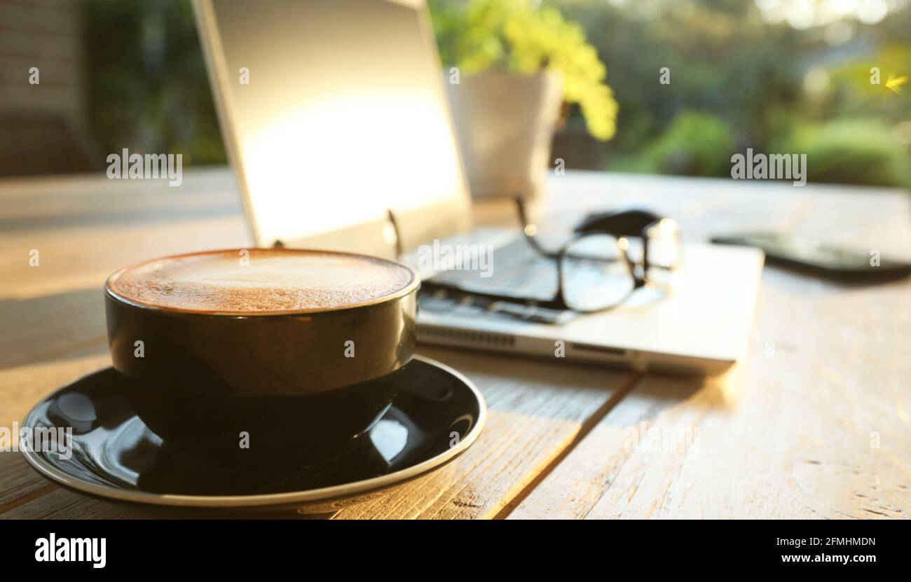 Bella mattina sole dorato su un tavolo esterno con caffè caldo, un computer portatile, telefono cellulare, occhiali e mouse per online sh Foto Stock