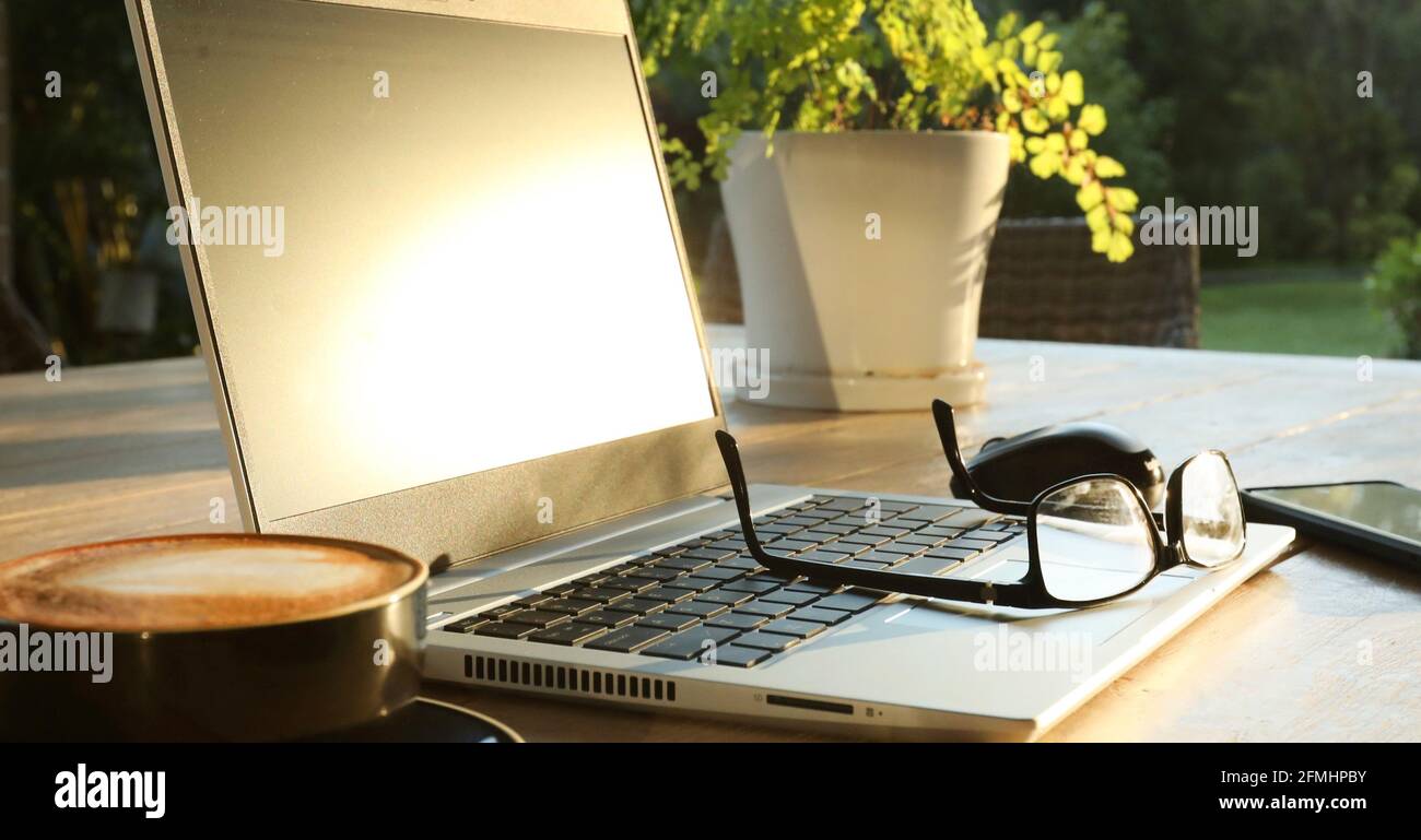 Bel sole dorato mattina su un tavolo esterno con caffè caldo, un computer portatile, cellulare, bicchieri e mouse pronti per onlin Foto Stock