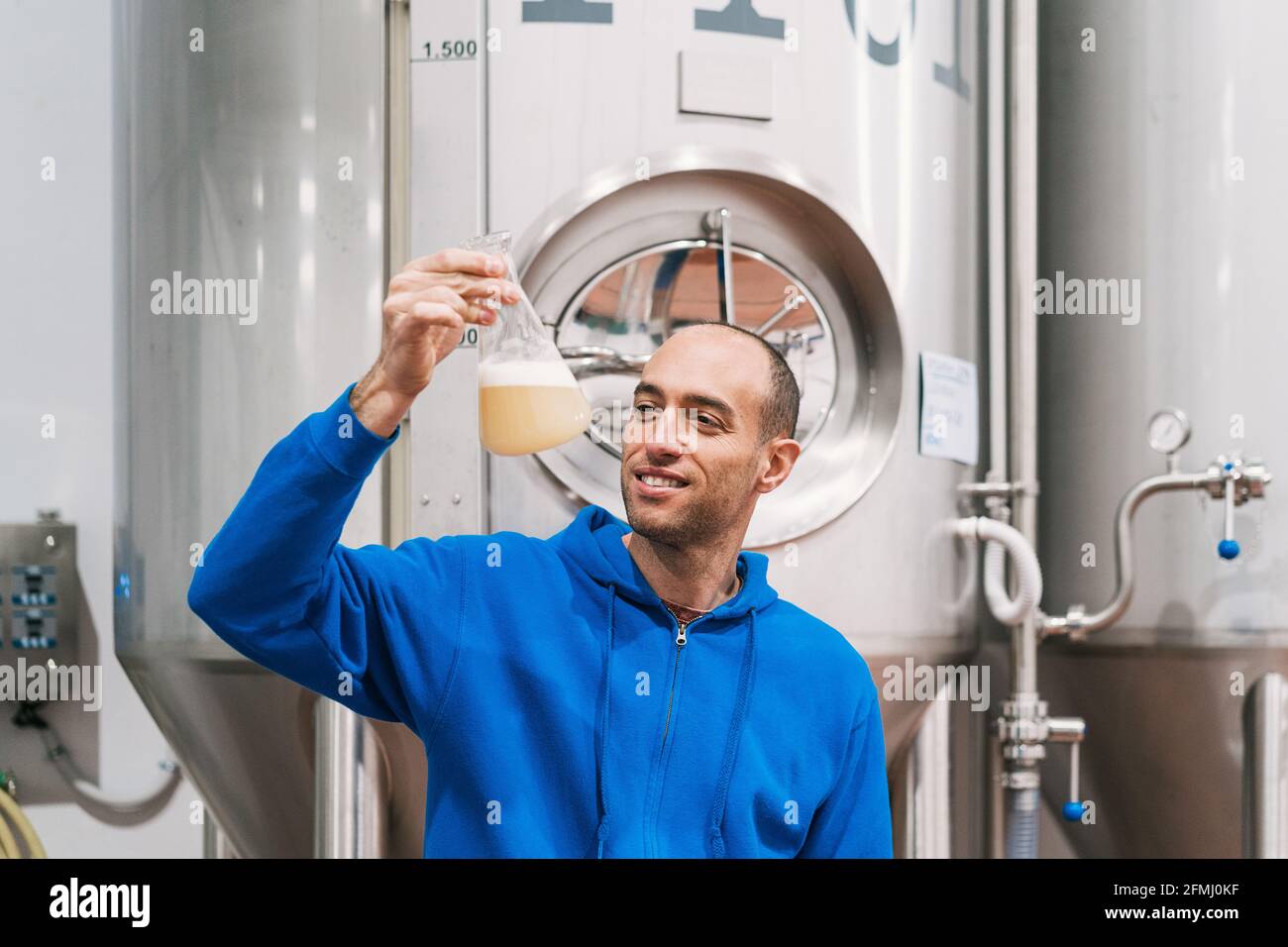 Allegro imprenditore maschio con liquido alcolico in fiasca che lavora contro vasche di fermentazione in birreria Foto Stock