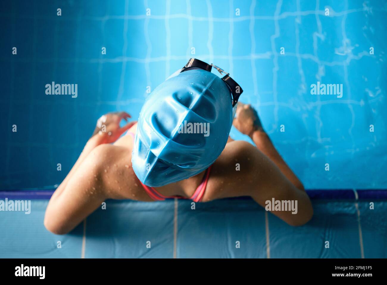 Vista dall'alto di uno sportivo anonimo in cuffia da nuoto appoggiata con le mani a bordo piscina durante la pausa dall'allenamento Foto Stock