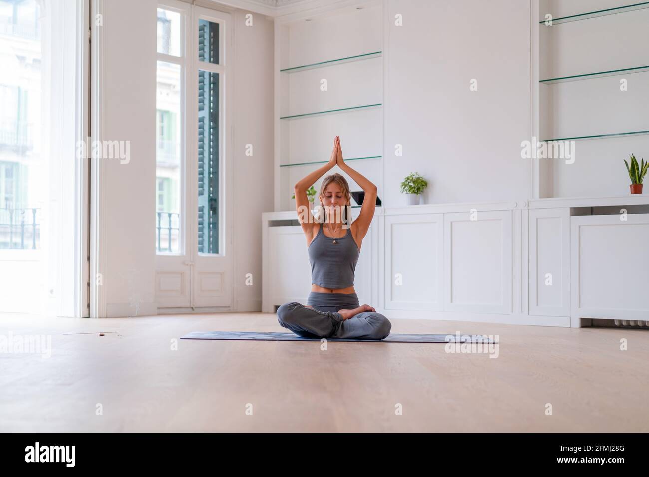 Donna tranquilla in activewear seduta in Lotus posa con la preghiera mani e mediare con gli occhi chiusi mentre si pratica yoga su tappetino in camera Foto Stock