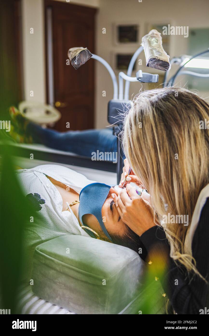 Coltivare cosmetologist irriconoscibile con pinzette che applicano ciglia false per l'estensione sull'occhio del cliente etnico con maschera protettiva del viso in salone durin Foto Stock