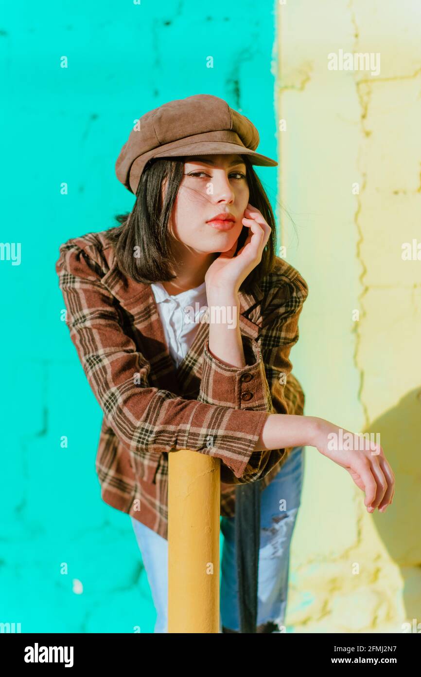 Giovane donna etnica in abbigliamento alla moda che tocca il viso mentre si inclina in avanti e guardando la fotocamera alla luce del sole Foto Stock