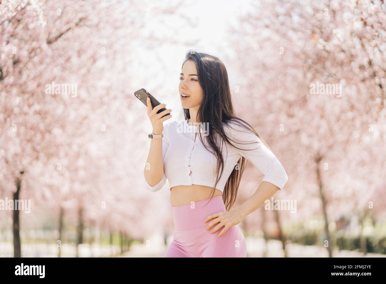 Giovane tenera femmina con mano sull'anca inviando un messaggio vocale sul cellulare mentre si guarda via alla luce del giorno Foto Stock