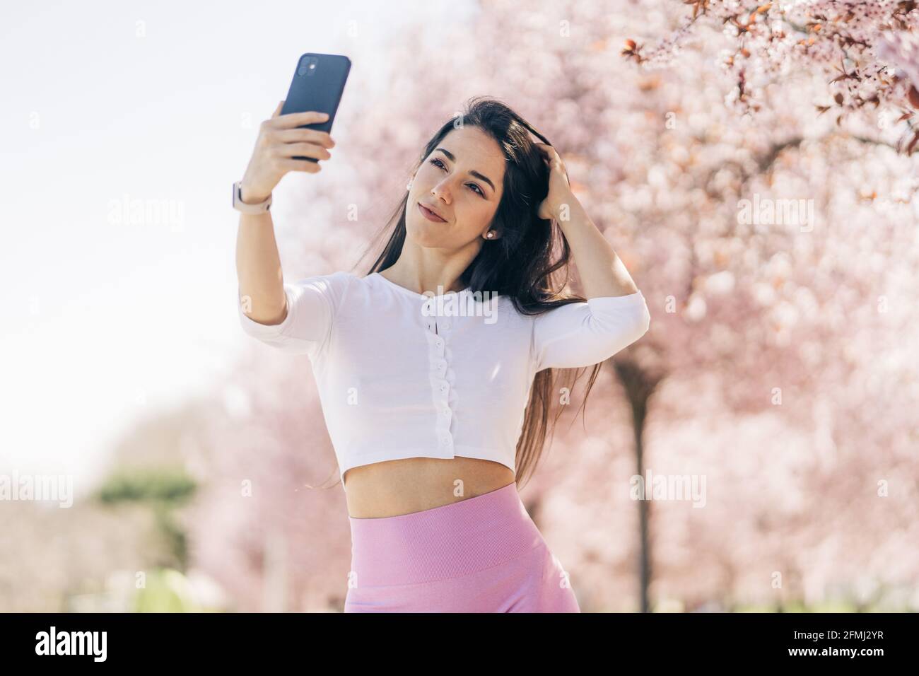 Giovane tenera femmina che prende selfie con il cellulare mentre tocca i capelli di luce diurna Foto Stock