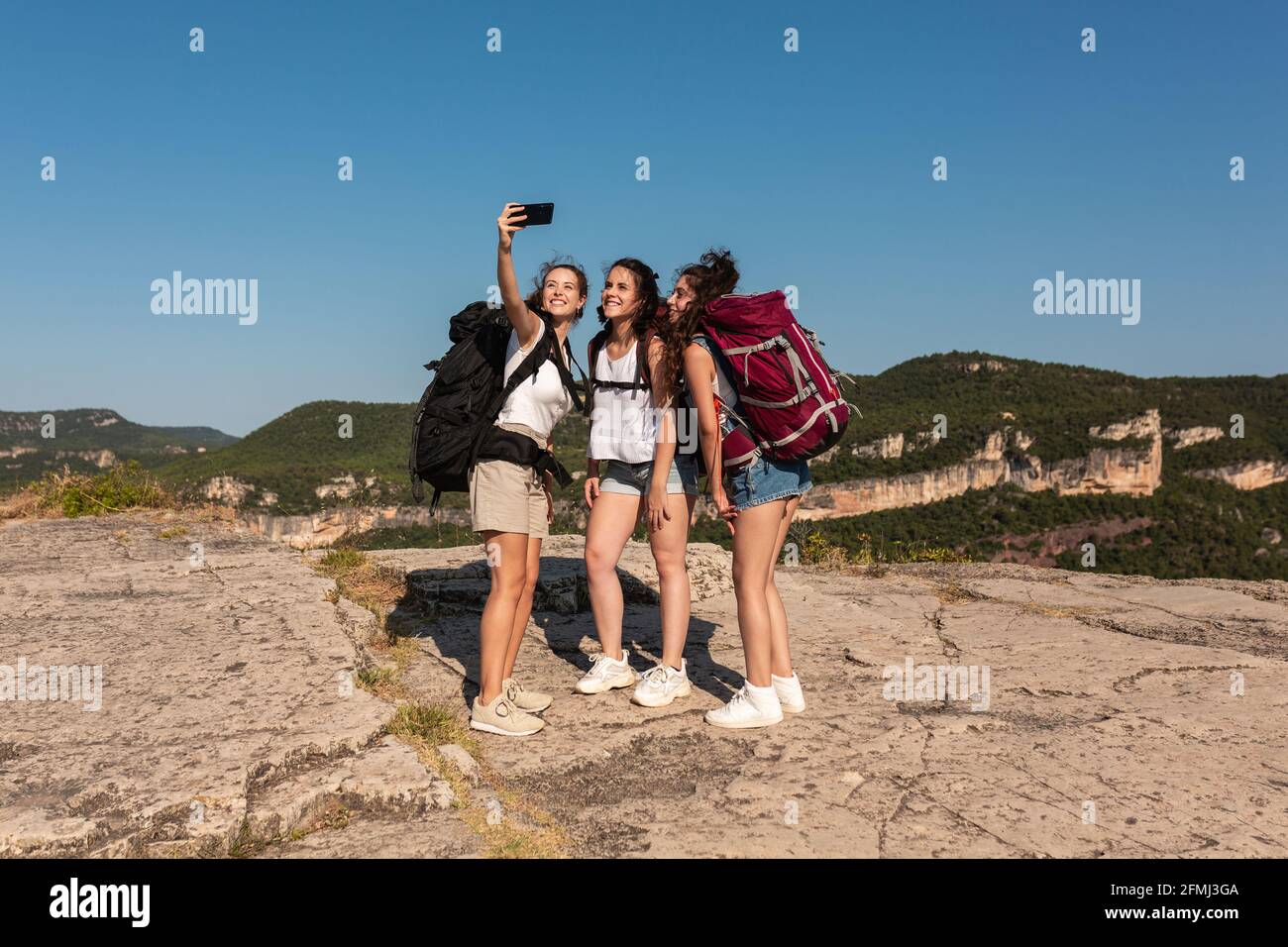 Compagnia di allegra backpackers femmina in piedi su collina in altipiani e di scattare da soli sullo smartphone durante le escursioni in estate Foto Stock