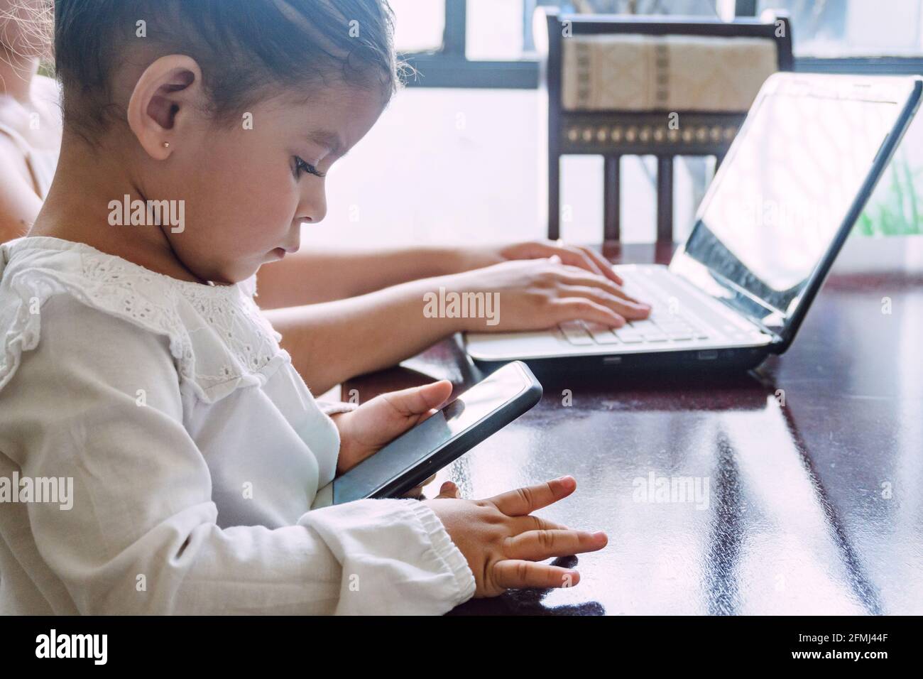 Vista laterale di etnico bambina che naviga telefono cellulare mentre seduto al tavolo con la madre di raccolto che scrive sul laptop a. casa Foto Stock