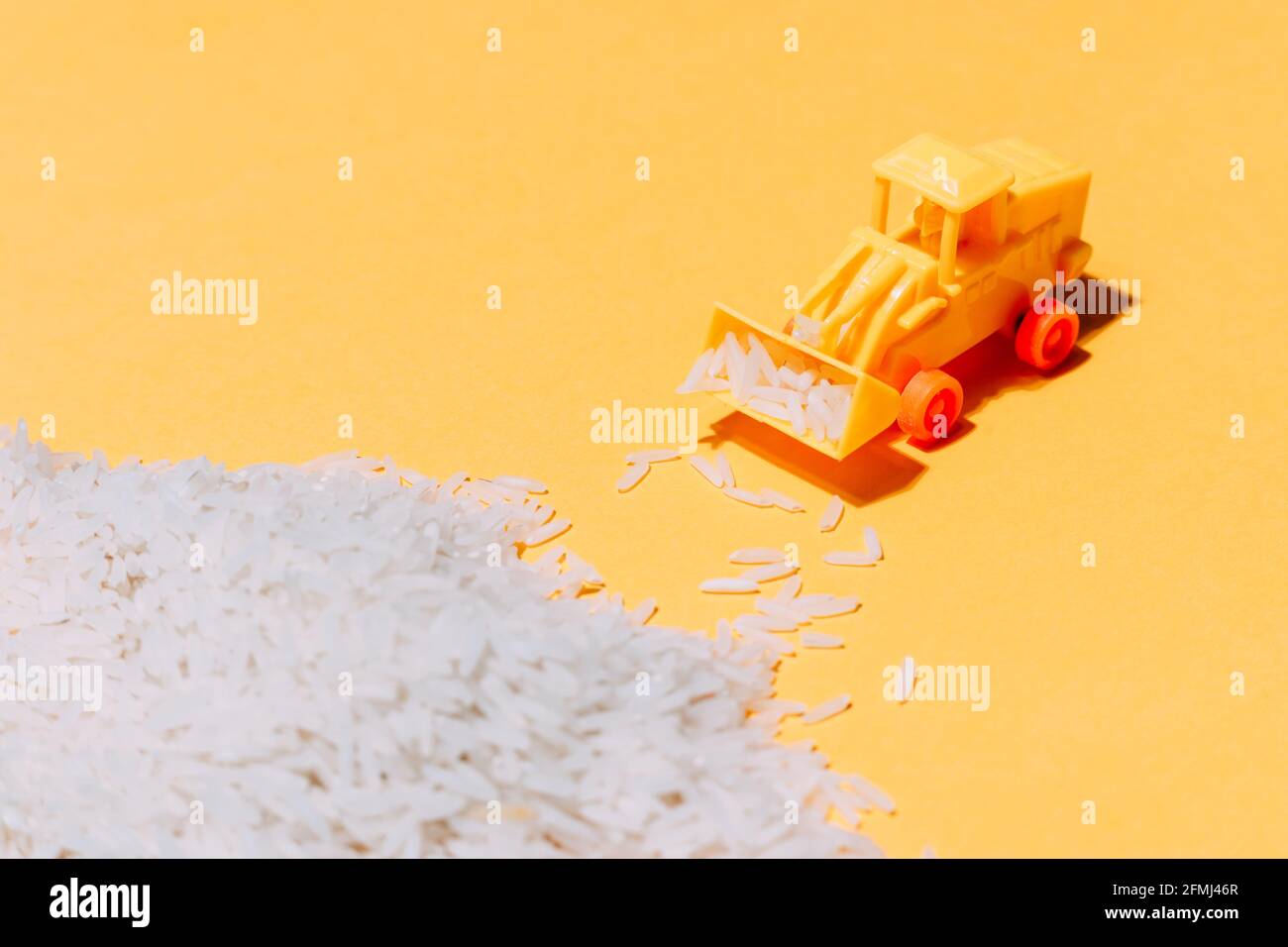 Dall'alto di un piccolo trattore giocattolo di plastica che raccoglie pile di grani di riso sparsi su sfondo giallo Foto Stock
