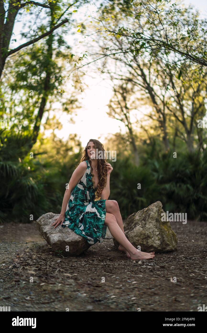 Pieno contenuto corpo giovane femmina che indossa trendy maxi sundress toccare capelli delicatamente e guardando via con il sorriso mentre si siede sopra grande pietra in sole verdeggiante Foto Stock