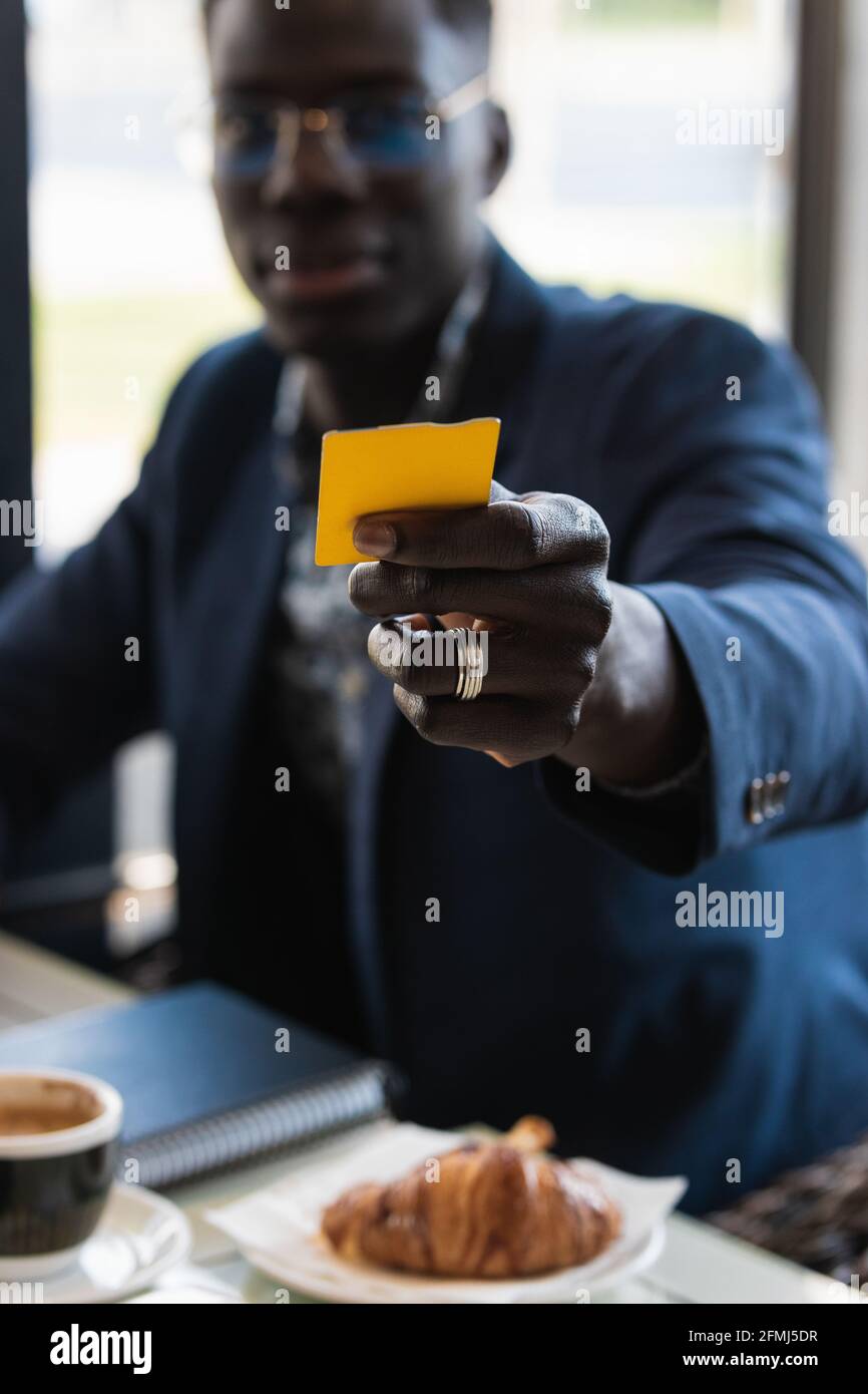 Crop uomo d'affari afro-americano con biglietto da visita seduto alla caffetteria tavolo durante la colazione e guardando la macchina fotografica Foto Stock