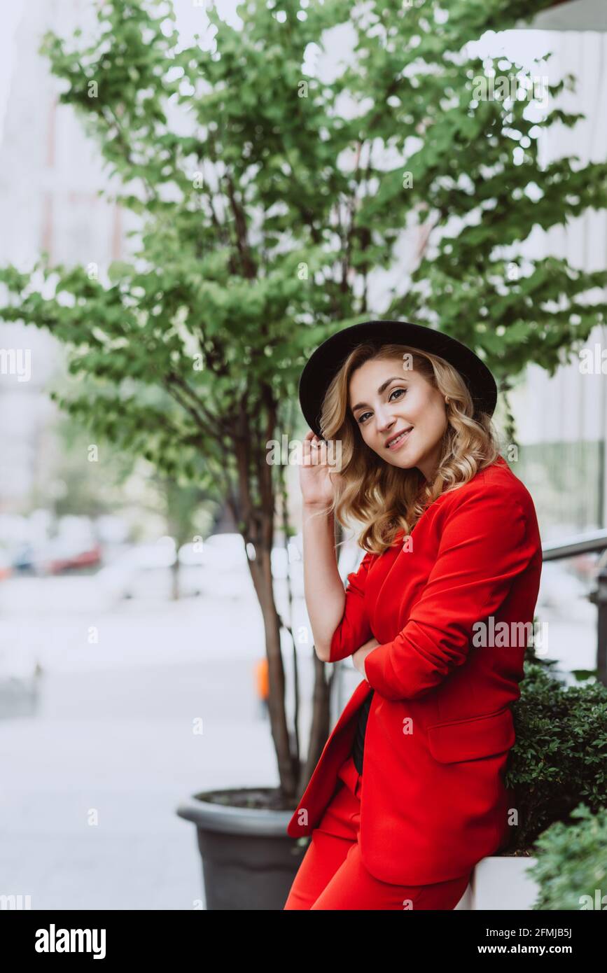Giovane bella donna bionda in un vestito rosso e cappello nero si erge all'aperto nel cortile accanto all'edificio. Persone eleganti. Messa a fuoco selettiva morbida. Foto Stock
