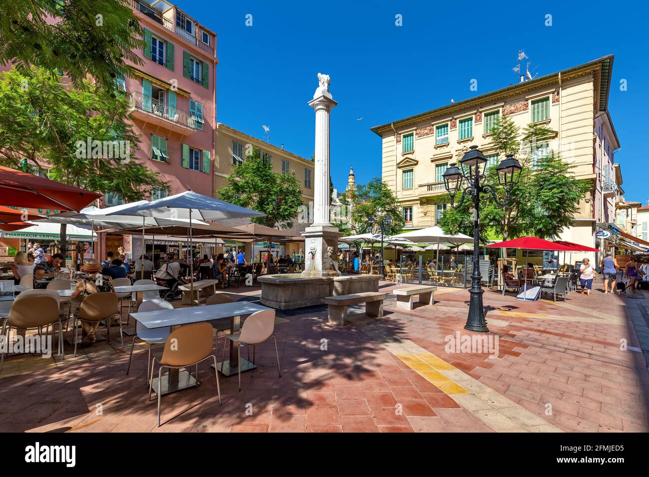 Persone sedute in ristoranti all'aperto sulla piccola piazza della città tra un bellissimo edificio sotto il cielo blu a Mentone, Francia. Foto Stock