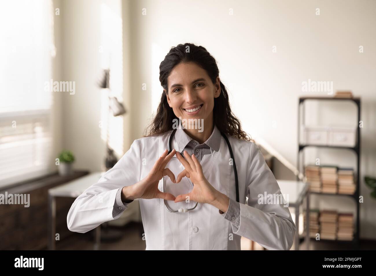 Una cardiologa latina sorridente guarda la fotocamera mostrare il cuore del dito Foto Stock