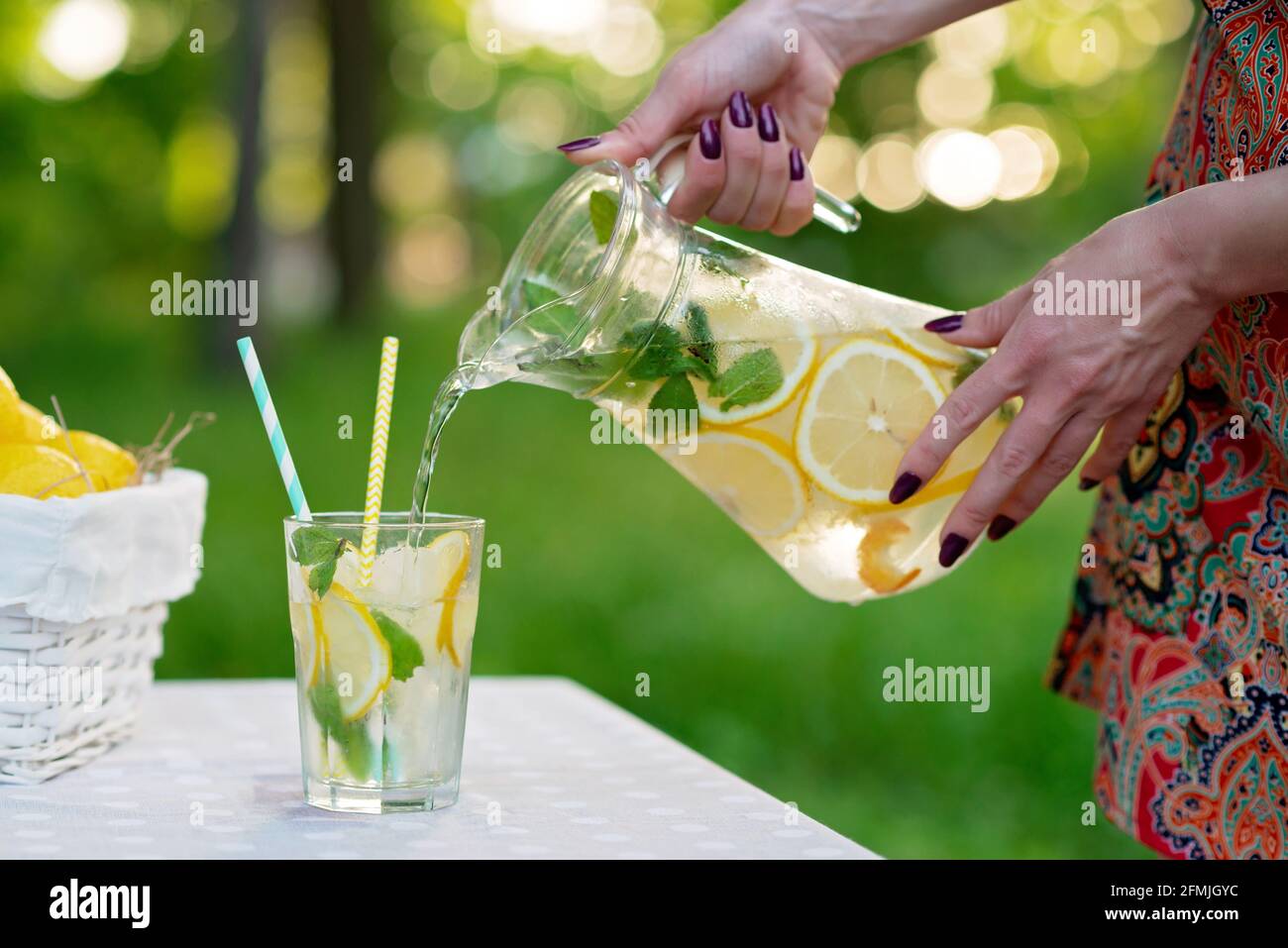 Le mani femminili versano una rinfrescante limonata fredda con i limoni nel ghiaccio e la menta da una caraffa in un bicchiere su un tavolo da giardino estivo su un picnic. Soft selective fo Foto Stock