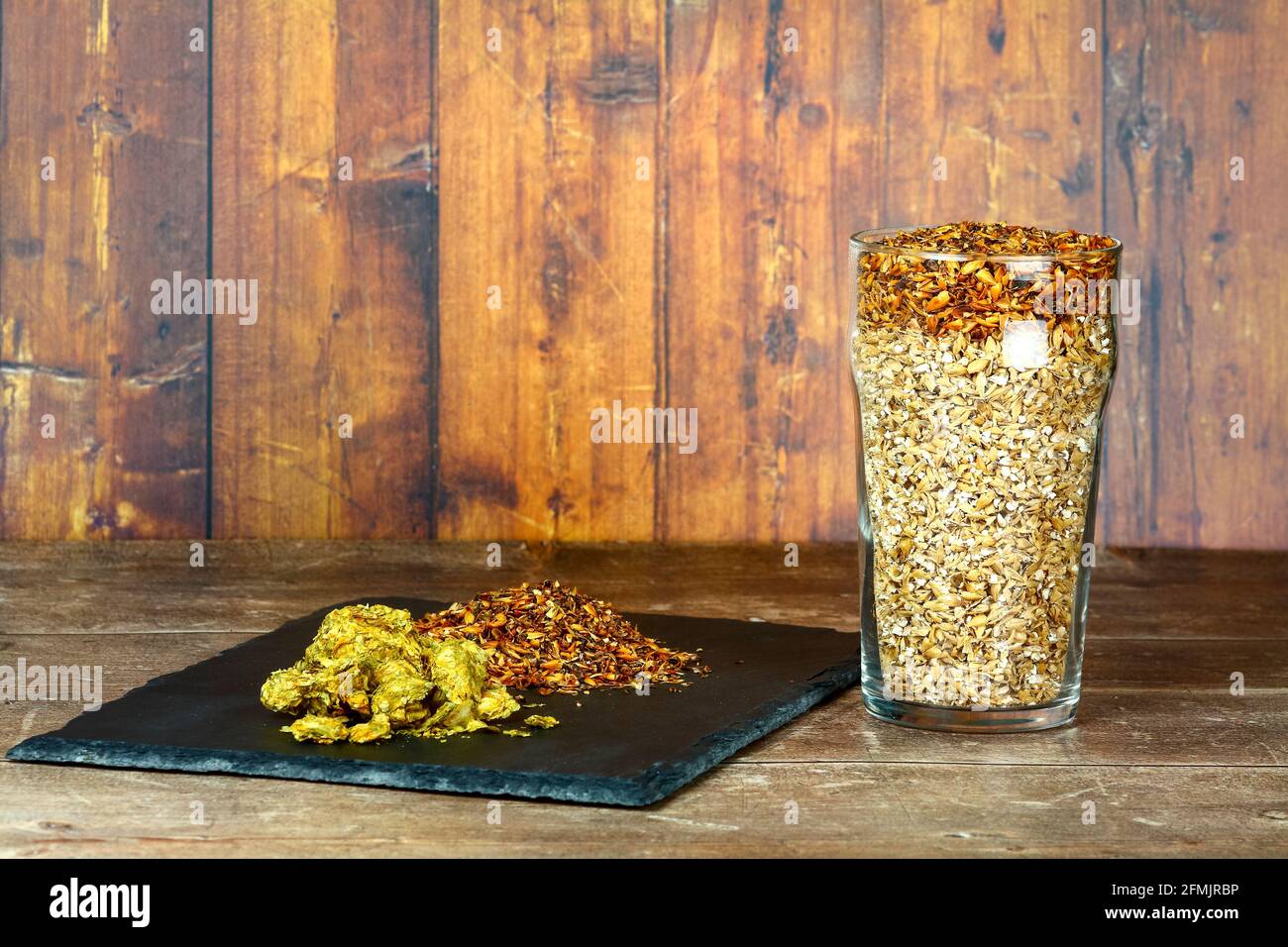 Vetro di pinta riempito con malto d'orzo gli ingredienti crudi utilizzati per fare le birre artigianali con luppolo su un'ardesia serving piatto Foto Stock