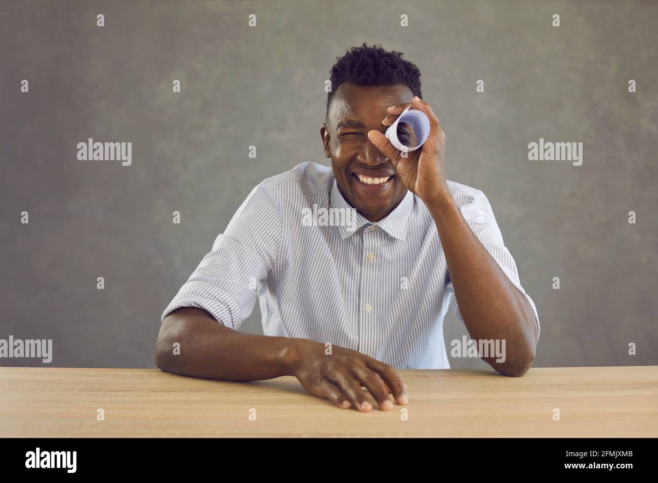 Uomo nero felice seduto alla scrivania e guardando attraverso il buco in rotolo di carta come nel telescopio Foto Stock