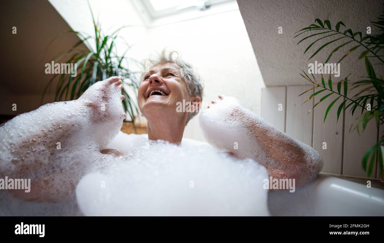Donna anziana allegra che si lava nella vasca da bagno a bolle a casa, ridendo. Foto Stock