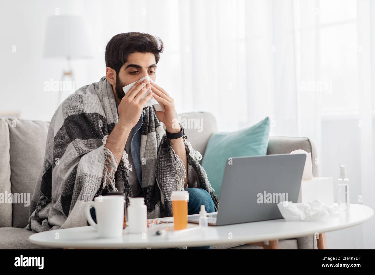 Uomo arabo malato coperto di coperta calda guardando lo schermo del computer portatile e starnutire il naso in carta tissue, seduto sul divano a casa, spazio vuoto. Sinusite illne Foto Stock
