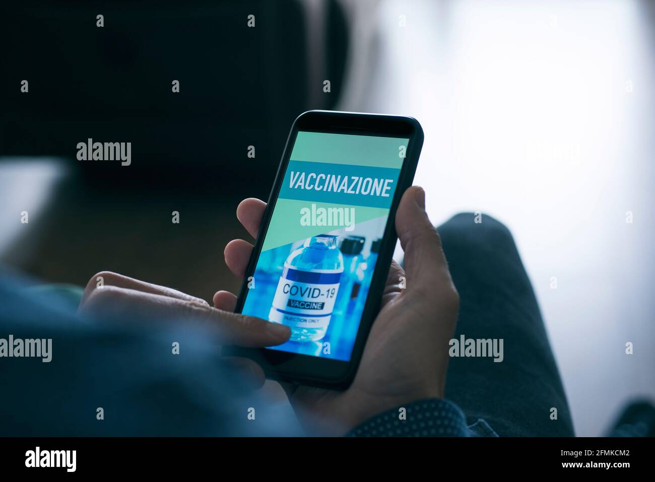 un giovane caucasico, seduto in soggiorno, guarda il suo smartphone, che legge il testo della vaccinazione scritto in italiano sul suo schermo Foto Stock
