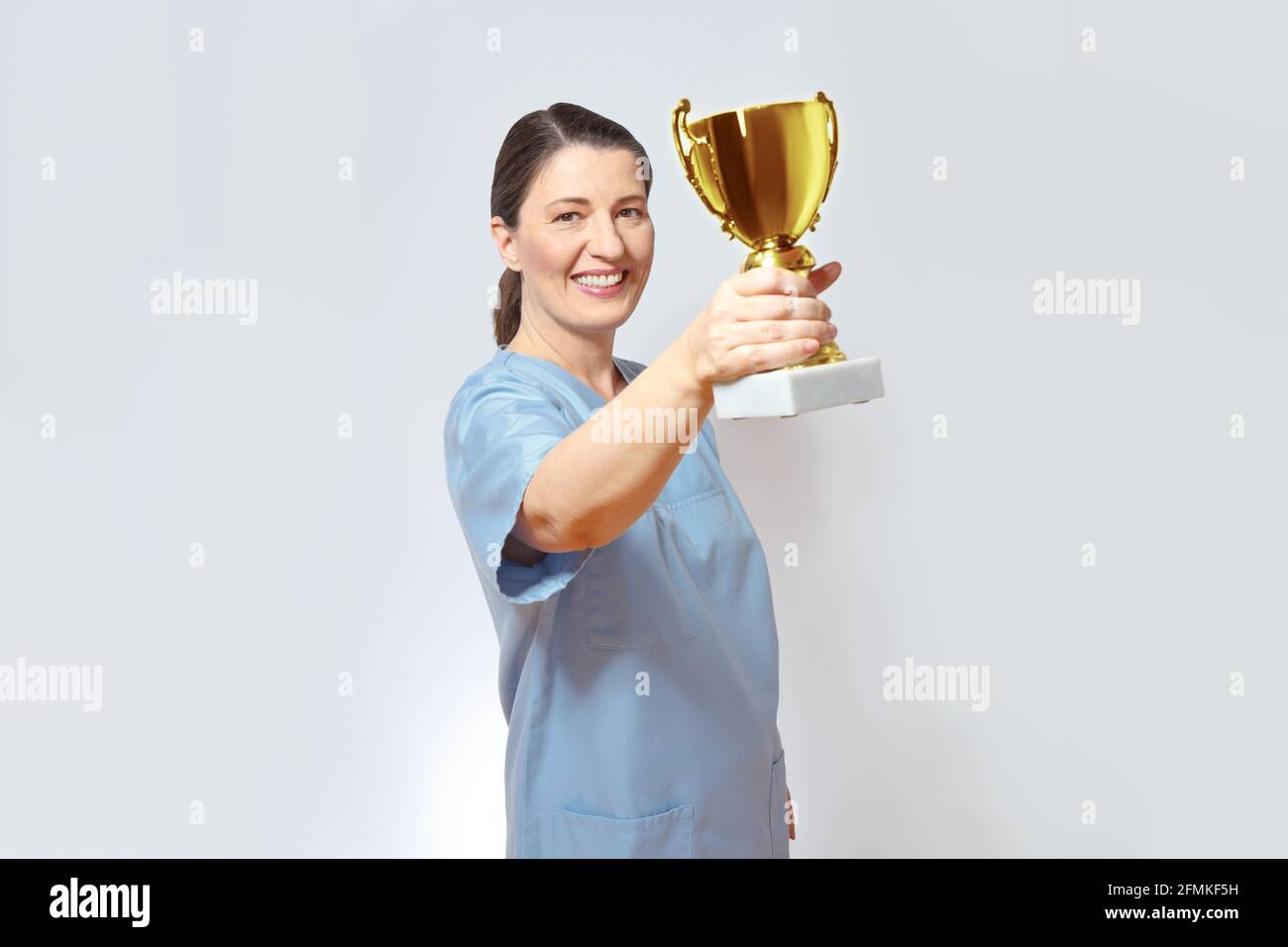Infermiera di media età in uno scrub azzurro sorridente orgogliosamente e tenendo un trofeo d'oro su, sfondo bianco, spazio di copia. Foto Stock