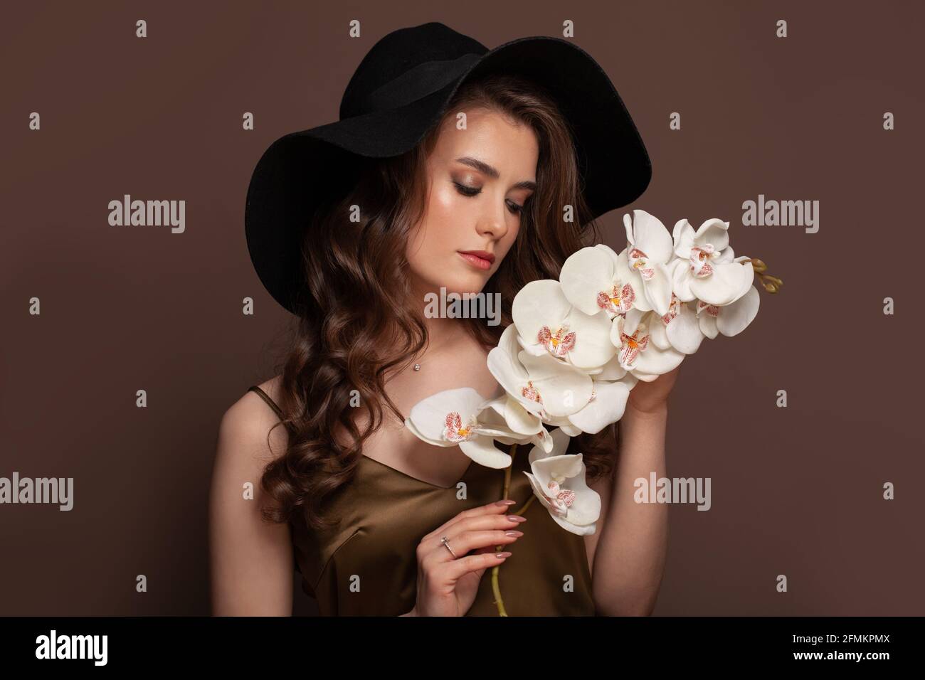 Bella donna con lunghi capelli ricci sani e scuri in nero cappello con fiori bianchi su sfondo marrone Foto Stock