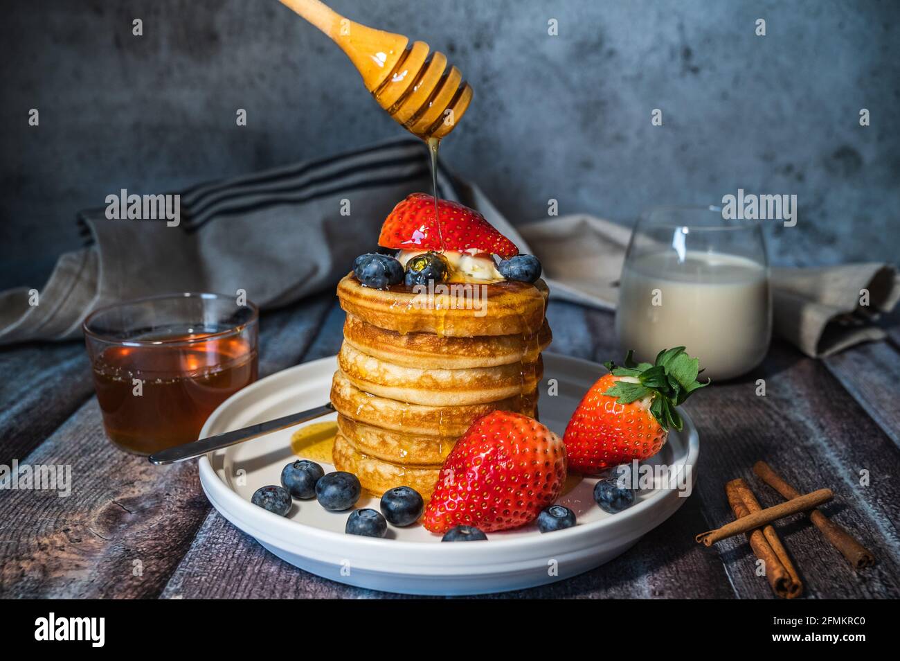 Una pila di frittelle soffici e soffici con sciroppo d'acero versato da bastone, mirtillo fresco, fragola e un bicchiere di latte su tavola di legno, delizioso essiccatoi Foto Stock