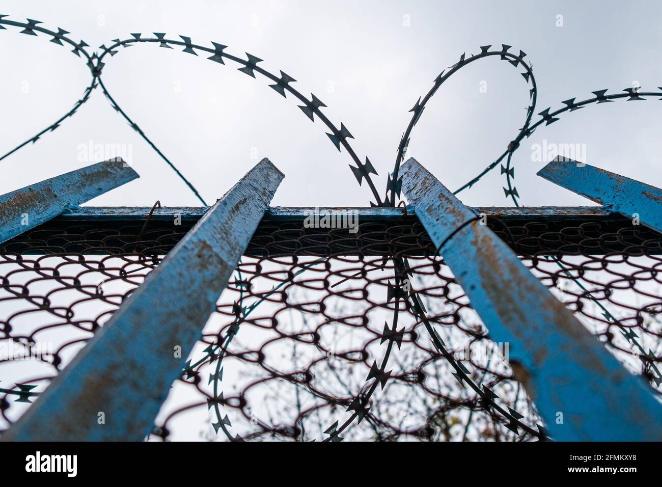 Una recinzione blu con filo spinato vicino alla prigione o all'ospedale mentale sullo sfondo del cielo grigio. Foto Stock